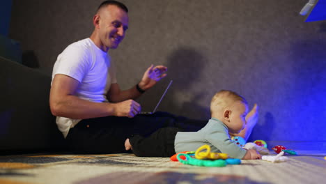 tiny little boy lies on the floor looking at his toys. dad sitting behind turns the kid his face to camera.