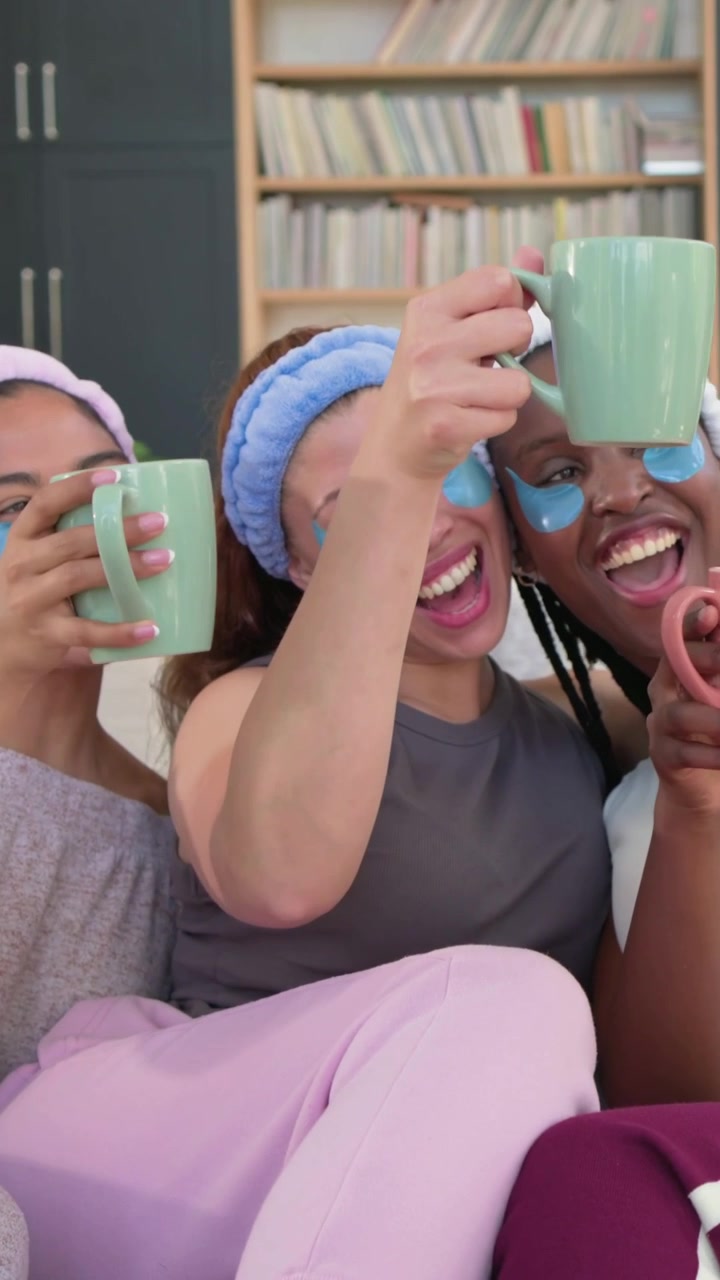 Vertical video: Giggling 3 women in plush headbands, gels on couch toasting for spa checking phone