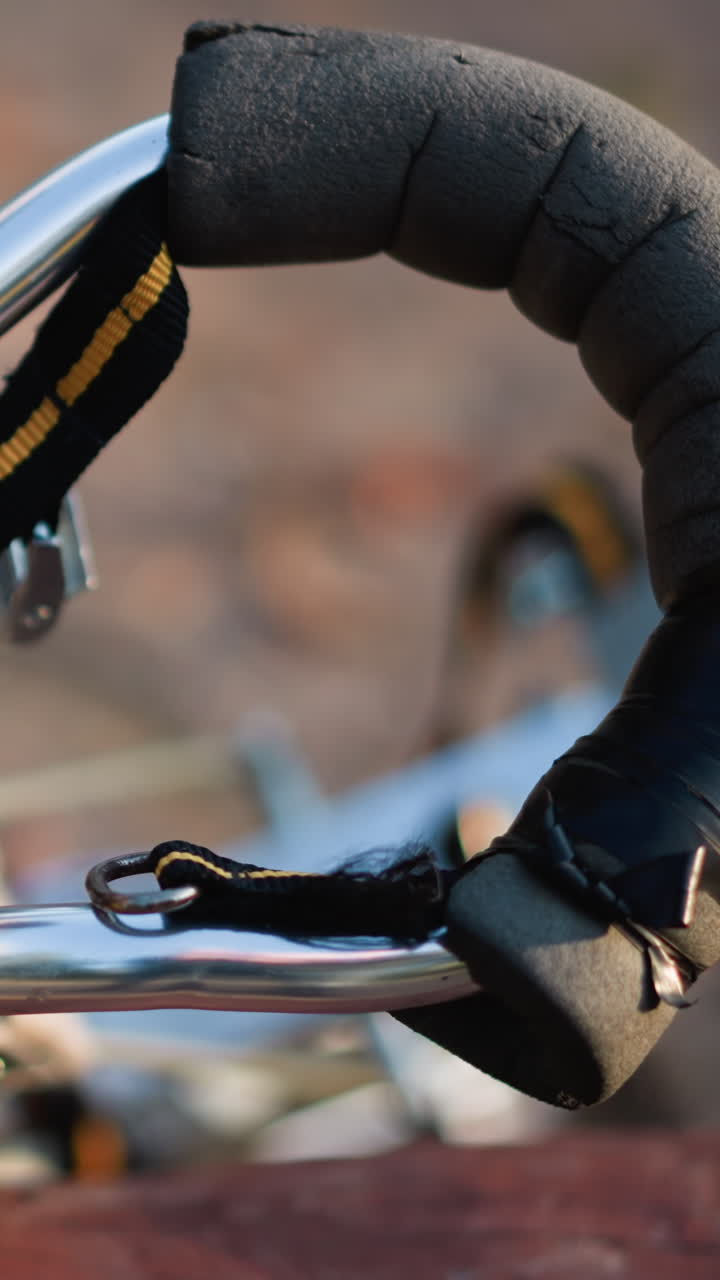 cerrado de pilar de metal con correa negra y naranja, mango envuelto con cinta negra y superficie metálica reflectante, posicionado al aire libre sobre un fondo borroso, resaltando detalles del equipo