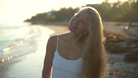 langzame beweging van volwassen vrouw achtergrondverlicht op het strand zwaait haar haar rond van zijde tot zijde uiteindelijk kijken en glimlachen naar de camera