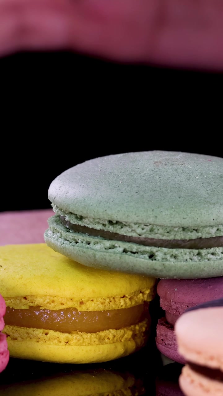A hand carefully arranges assorted pastel macarons on a glossy black surface under bright, even lighting. Close-up, steady camera highlights dessert textures and vibrant colors