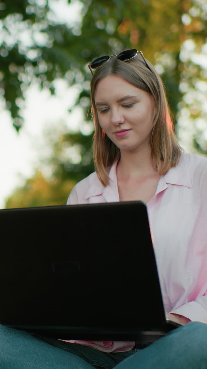 joven sentada con las piernas cruzadas en un campo de hierba, trabajando en una computadora portátil, mira hacia arriba pensativa con una cálida sonrisa, el fondo presenta árboles y vegetación, reflejando la calma y la productividad al aire libre