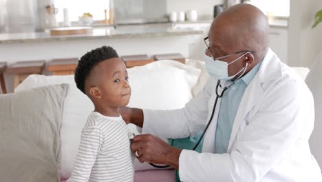 Senior-african-american-male-doctor-in-face-mask-using-stethoscope-on-boy-patient,-slow-motion