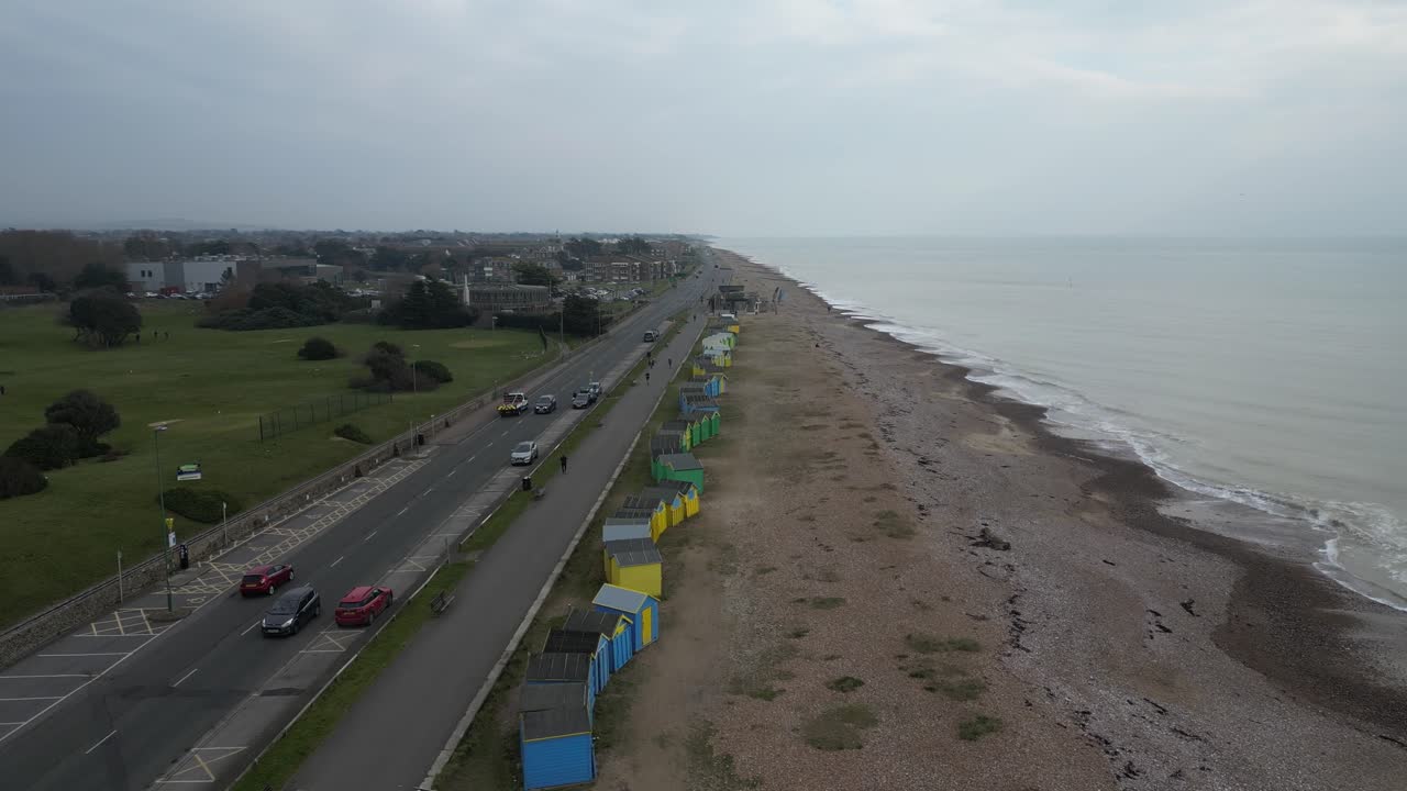 Aerial view of Brighton seafront on a cloudy day, featuring colorful beach huts, a long pebbled shoreline, and traffic along the coastal road beside a green open park space.