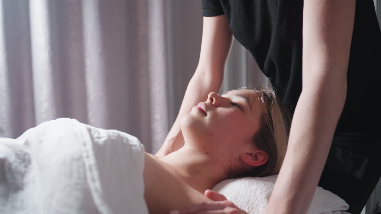 Light skin blond woman lying under white blanket while receiving chest massage from therapist dressed in black, captured in peaceful spa setting with natural light reflecting off curtain background