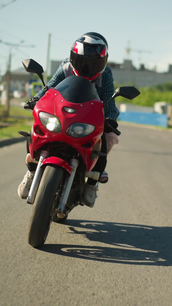 dos hermanas, ambas con cascos, montan una bicicleta eléctrica roja mientras navegan por la carretera, se pueden ver coches viniendo por detrás, con un borrón de edificios de la ciudad y postes eléctricos a lo largo de la carretera