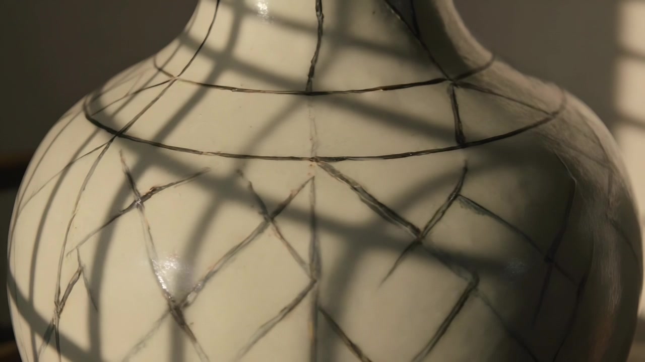 Telecamera inclinata che mostra un vaso color crema alla finestra, le lamelle della tenda che rivelano la smaltatura craquelé e il riverbero