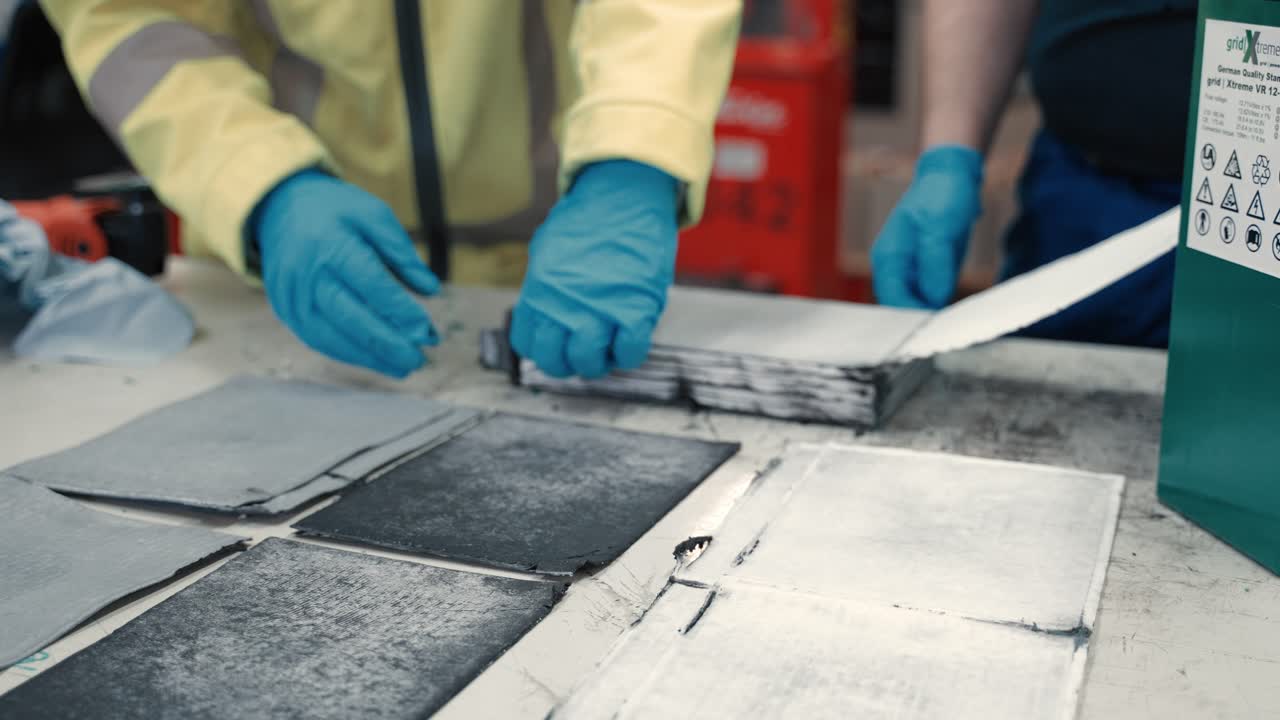 큰 배터리의 탄소 및 분리 계층이 두 명의 노동자들에 의해 테이블을 가로질러 놓이는 방법에 대해 자세히 살펴보십시오.