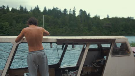 topless young male walks into frame scrapes old mastic from timber boat cabin roof planking