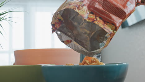 Close up of person hand returning caramel popcorn into packet on dark table with blue and green bowls nearby and blurred person passing by in bright indoor room with window light