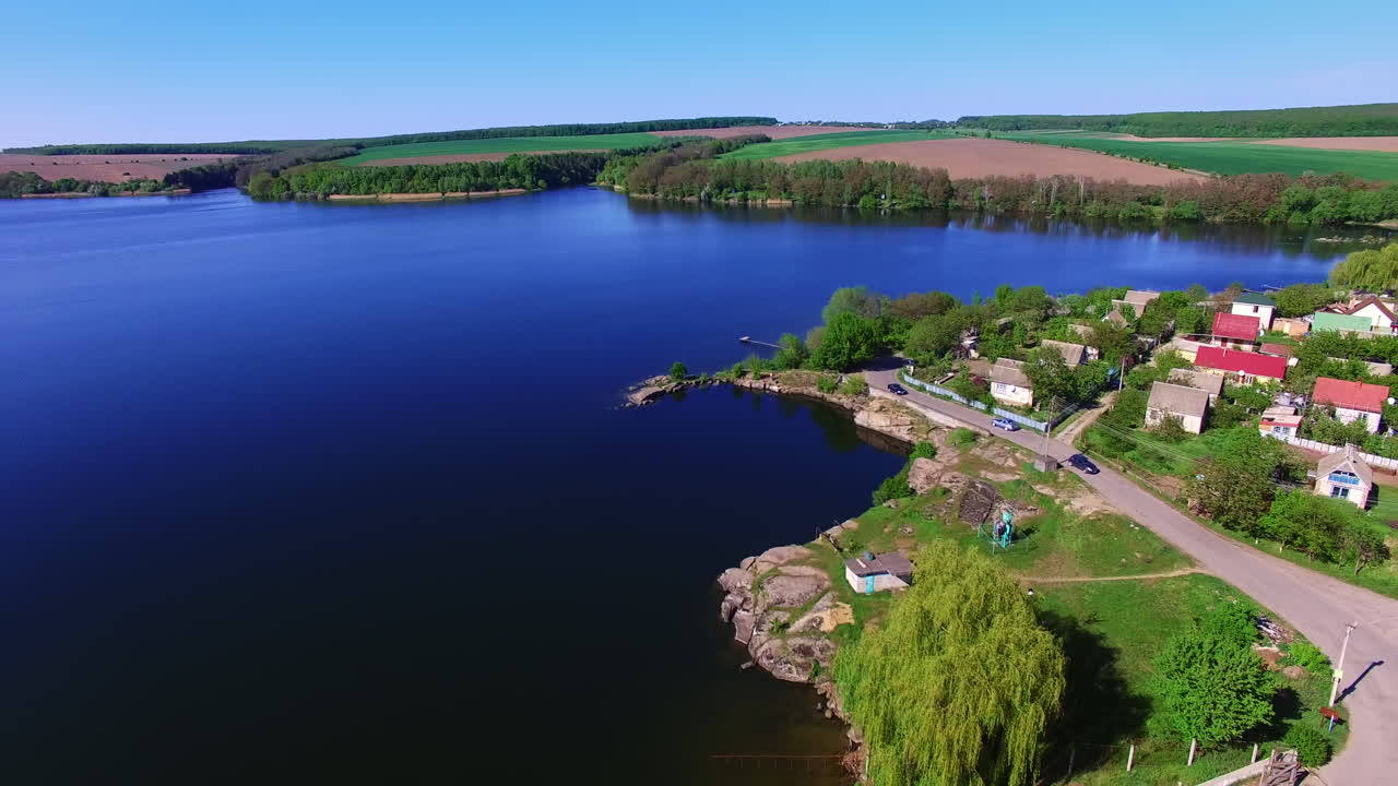 Automobile road with some cars at the waterfront. Drone flying over the azure river on sunny day. Agricultural fields at backdrop.
