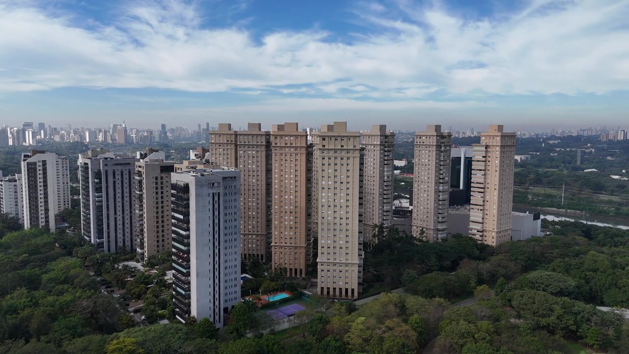 Tomada de drone orbital suave em torno de torres residenciais de alto padrão ao lado do exuberante Parque Villa-Lobos, na zona oeste de São Paulo, Brasil, exibindo arquitetura moderna, vegetação urbana e um céu azul claro.