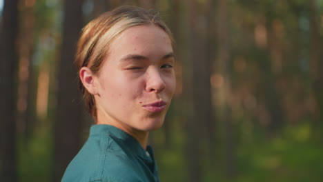 close-up of woman in green shirt standing outdoors in forest, turning her head with a subtle smile as she blows a playful kiss to the air, sunlight filters through trees