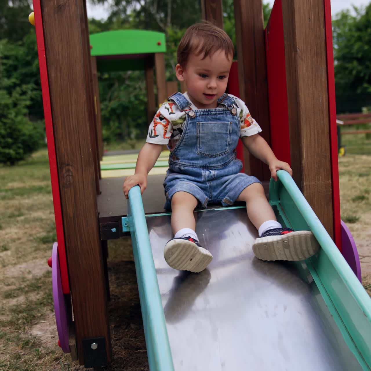 Beautiful baby boy in jeans romper sits on top of the slide. Kid hesitantly decides to go down smiling sweetly