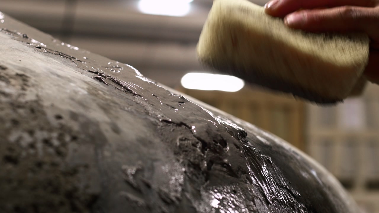 Follow shot with a slow tilt down as a hand uses a sponge to smooth wet grout on the curved surface of a pizza oven during construction.