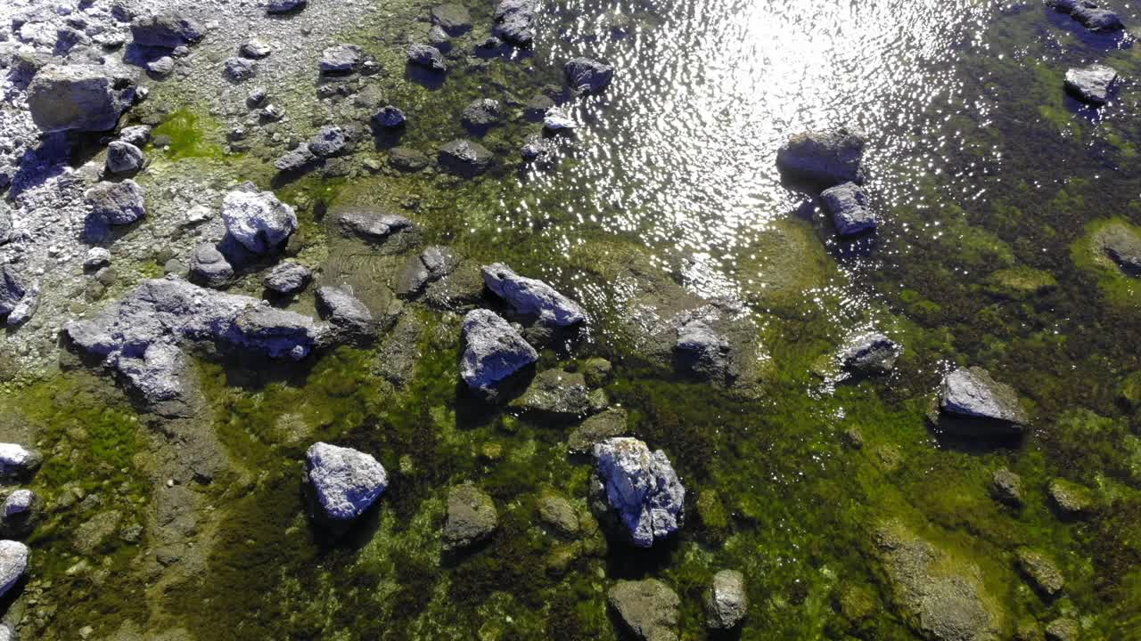 Aerial view over rocks, in shallow water, on the coast of Faro island, sun glittering on the water surface, on a warm, summer day, in Gotland, Sweden - low, overhead, drone shot