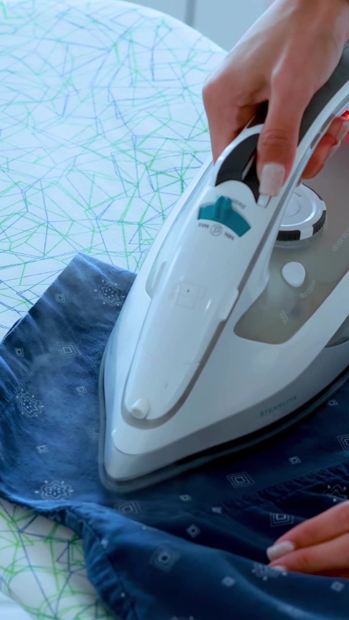 A person uses a steam iron to press a blue patterned shirt on a modern ironing board in bright, natural lighting. Close-up, steady shot