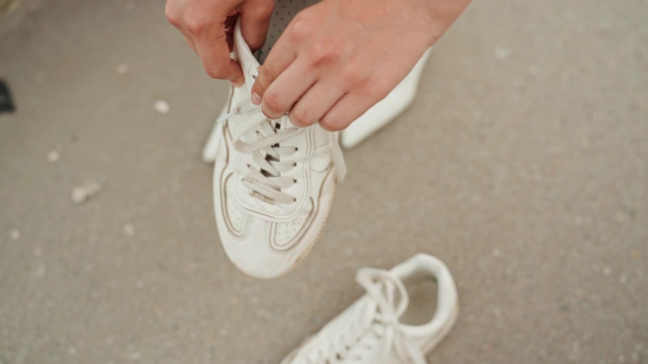 Una donna allaccia i lacci di una sneaker bianca, concentrandosi sulle mani e sui dettagli della scarpa. L'azione si svolge all'aperto con luce naturale che mostra un'attenta cura della calzatura.