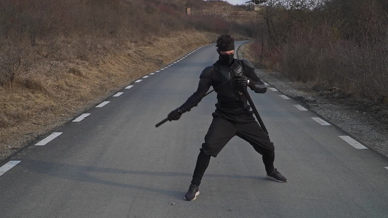 A masked martial artist dressed in black tactical gear is captured mid-motion while sheathing a katana on an empty rural road. The dramatic pose evokes a cinematic feel of discipline and warrior focus