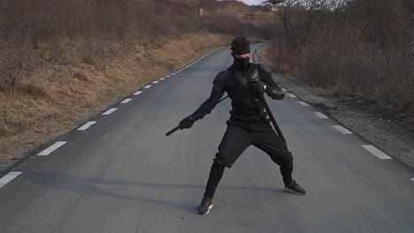 a masked martial artist dressed in black tactical gear is captured mid-motion while sheathing a katana on an empty rural road. the dramatic pose evokes a cinematic feel of discipline and warrior focus