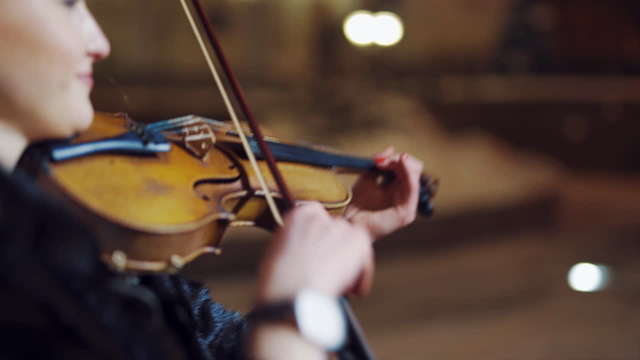 An attractive girl with long hair holds a violin in her hands and plays amazing melody on the background of high architecture in the city in the evening. Blurred background. Close-up