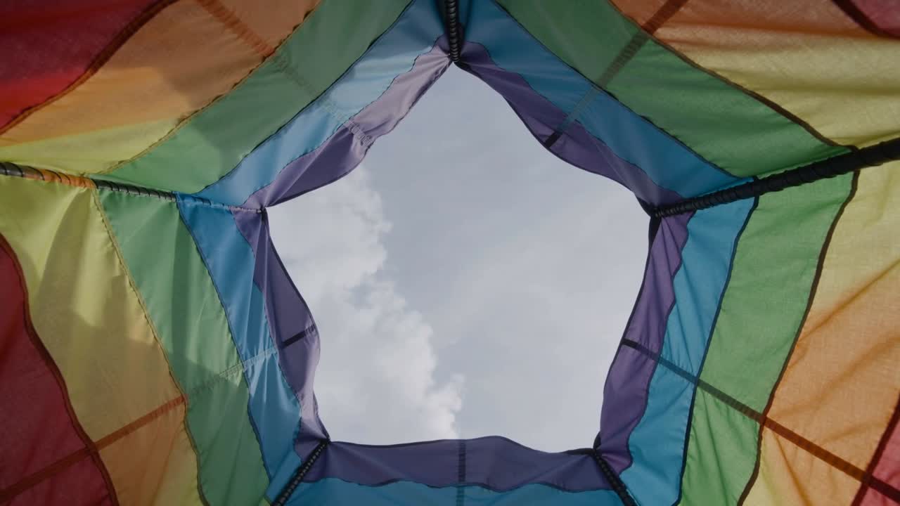Una brezza leggera che fa increspare una tenda esagonale multicolore sopra un parco, mostrando il cielo centrale