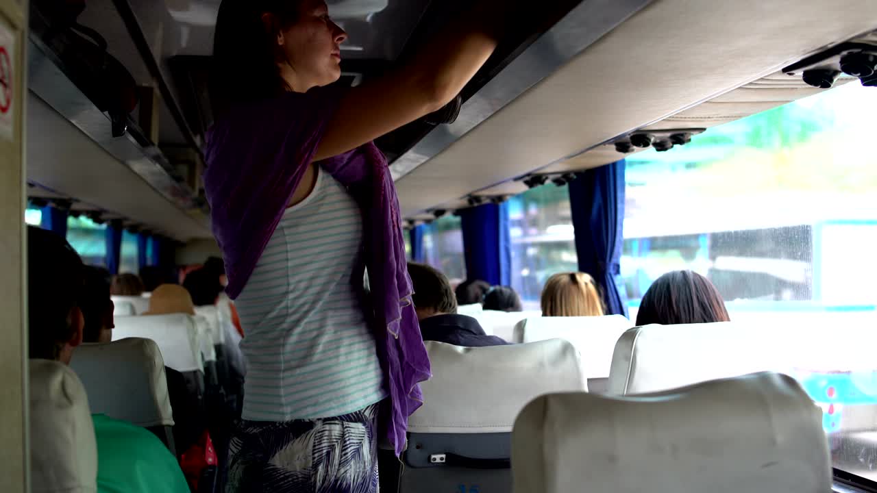 A woman walks through the cabin of the bus, removes the bag on the luggage rack and sits down