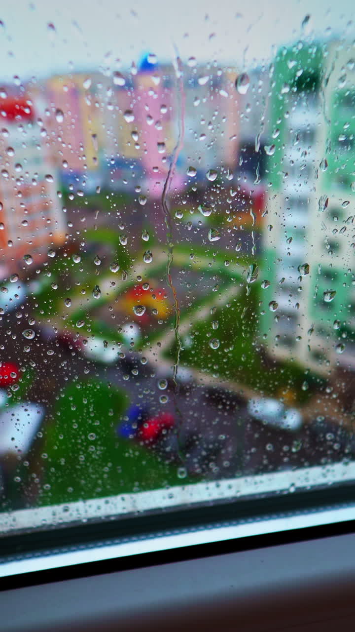 Rainy weather outside. View through the window with raindrops on glass on the background of colorful buildings in the city. Vertical video