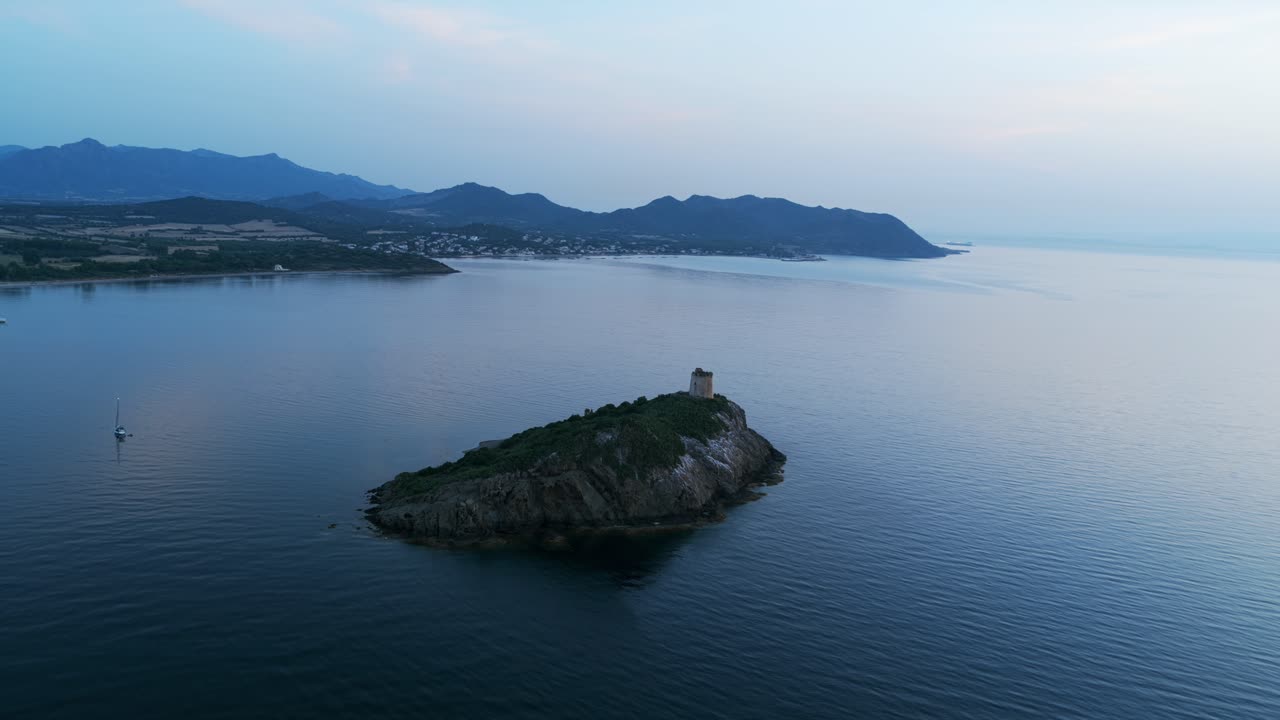 Historic watchtower on rocky islet near Pula, sailboat is anchored in calm sea at dusk with mountains in background, Italy. Aerial drone panoramic view, blue hour