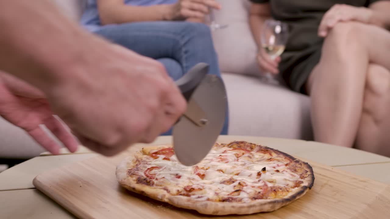 Primo piano di una mano che taglia una pizza fresca con un taglierino su un tagliere di legno mentre amici siedono su un divano sullo sfondo bevendo vino