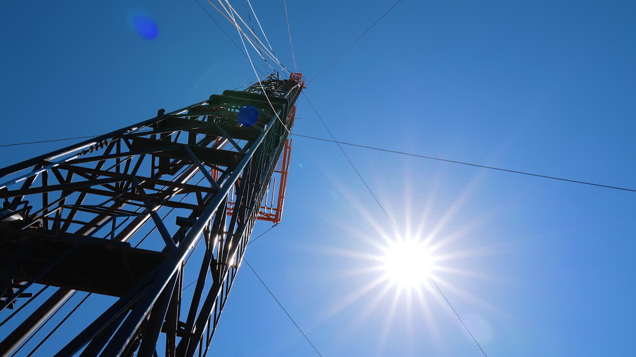 Derrick at the site for oil production. Low angle view at the drilling tower at the backdrop of blue sky with bright sun.