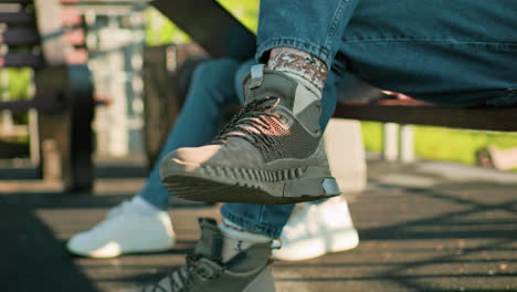 close-up of legs of two people seated outdoors on wooden benches, wearing casual sneakers and jeans, one with a laptop in sunlight casting shadows