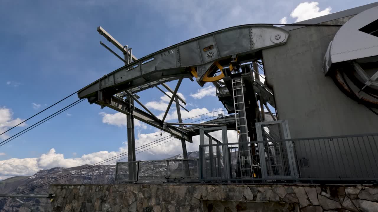 Una stazione della funivia si erge su una cima di montagna con cieli azzurri e sereni sopra. La struttura include cavi e una piattaforma. Cime innevate circondano la stazione, mostrando un paesaggio montano