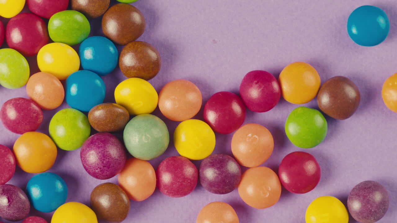 Colorful dragee candies on purple background. Woman hands moove bonbons. Summer bright texture studio shot for store,supermarket,close up top view. Children sweets made of sugar, mix of lollipops.