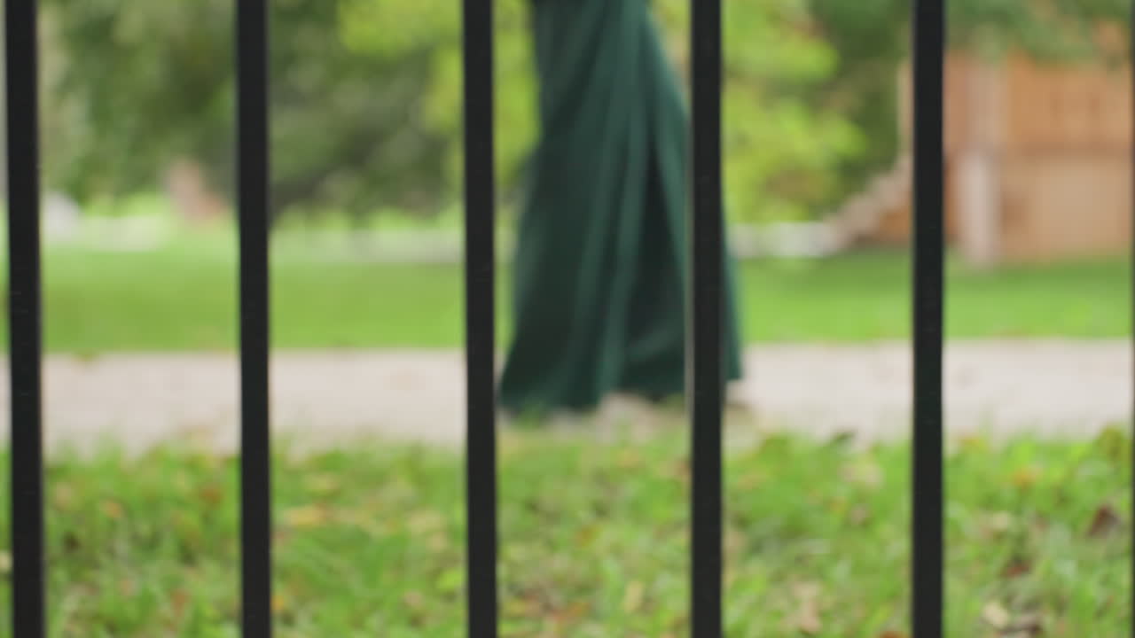 Gambe che camminano dietro una recinzione di ferro nel parco, sentiero, vialetto alberato e prato in una luce soffusa, barre verticali creano una prospettiva incorniciata, pantaloni casual e passo costante suggeriscono un pendolare riflessivo