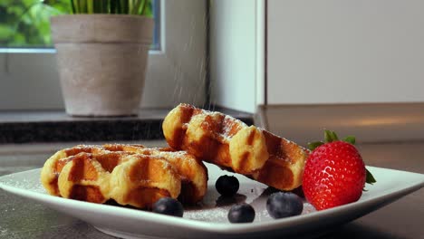 Static-shot-of-delicious-Belgian-waffles-decorated-with-fruity-blueberry-and-strawberry-while-sweet-powdered-sugar-is-added-in-the-kitchen