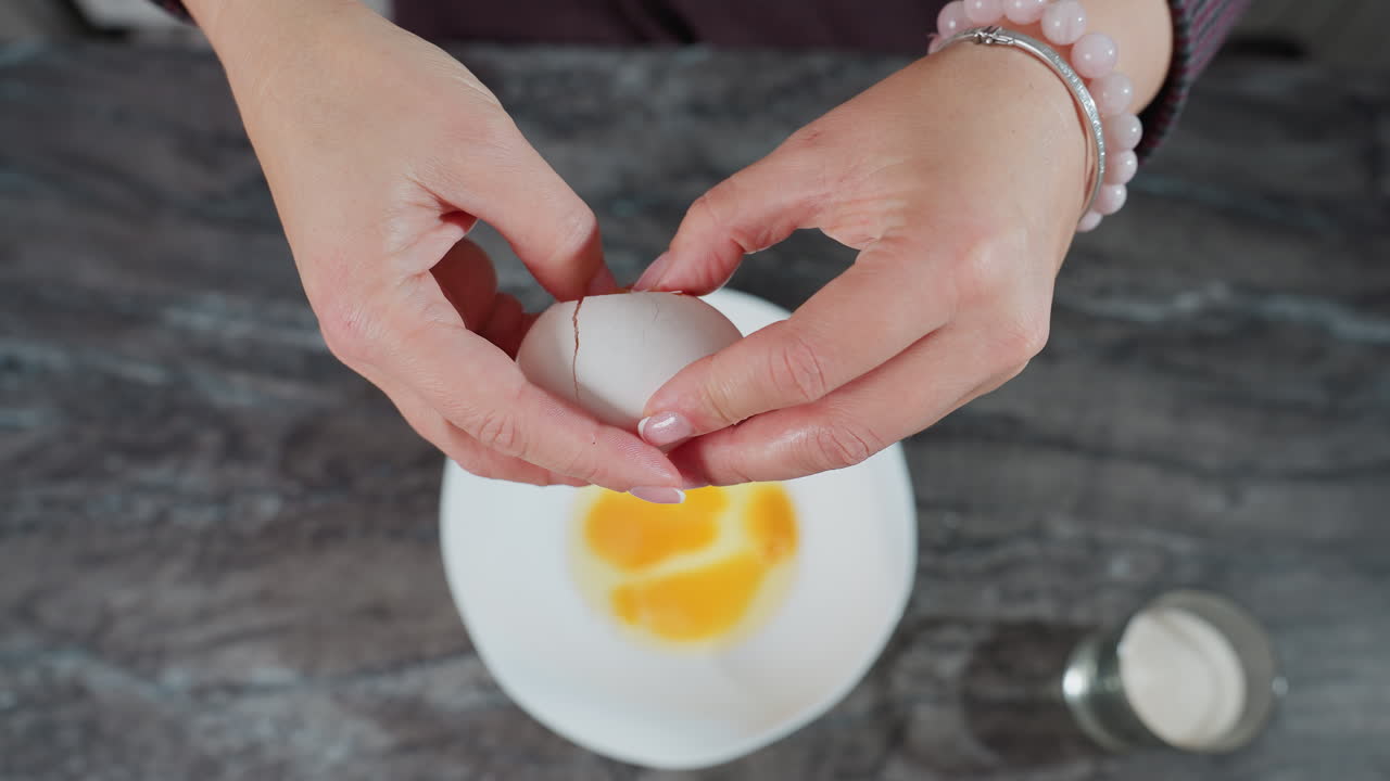 vista aérea de las manos del chef adornadas con una pulsera rompiendo un huevo en un cuenco blanco, las yemas de huevo son visibles dentro del cuenco, con una taza de azúcar cerca en una encimera de cocina oscura
