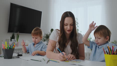 Una-Joven-Madre-Con-Dos-Hijos-Sentados-En-Una-Mesa-Blanca-Dibuja-Lápices-De-Colores-En-Papel-Ayudando-A-Hacer-La-Tarea