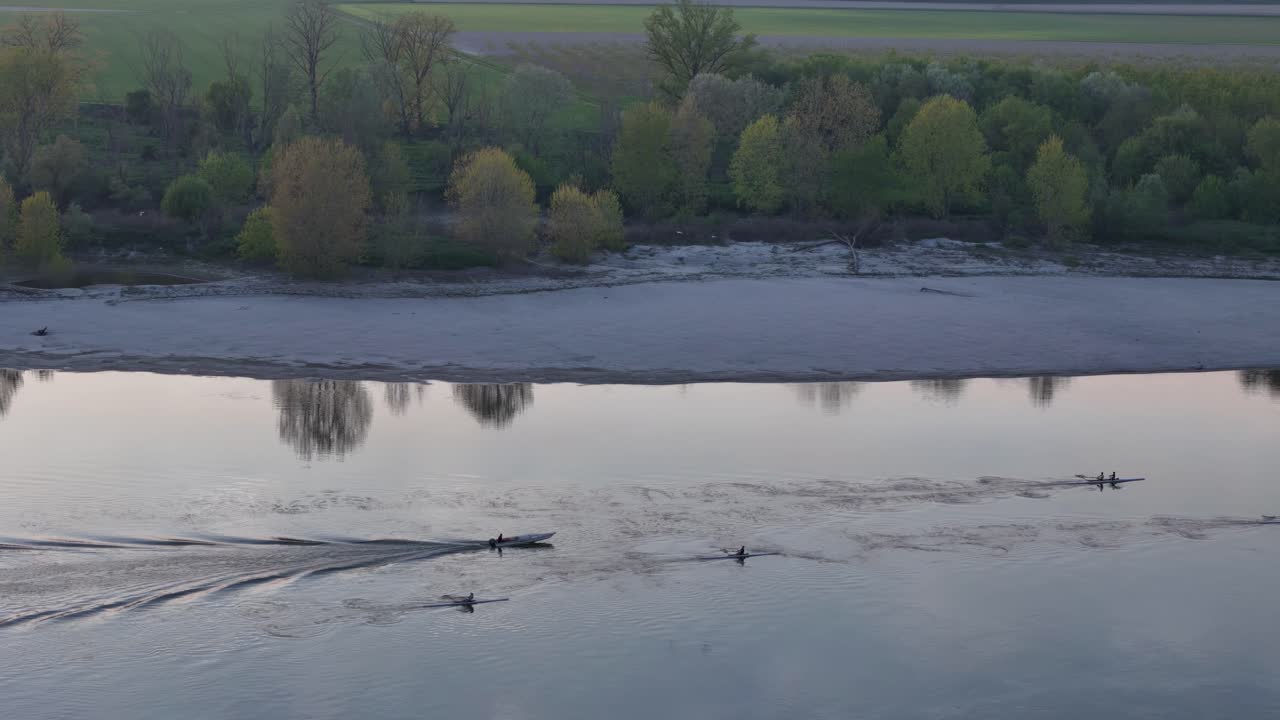 Real-time drone track captures rowing boats gliding across the Po River near Cremona at sunrise, trailing ripples through autumn reflections framed by floodplain trees and distant urban silhouettes