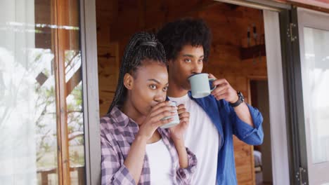 happy african american couple looking through window and drinking coffee in log cabin, slow motion