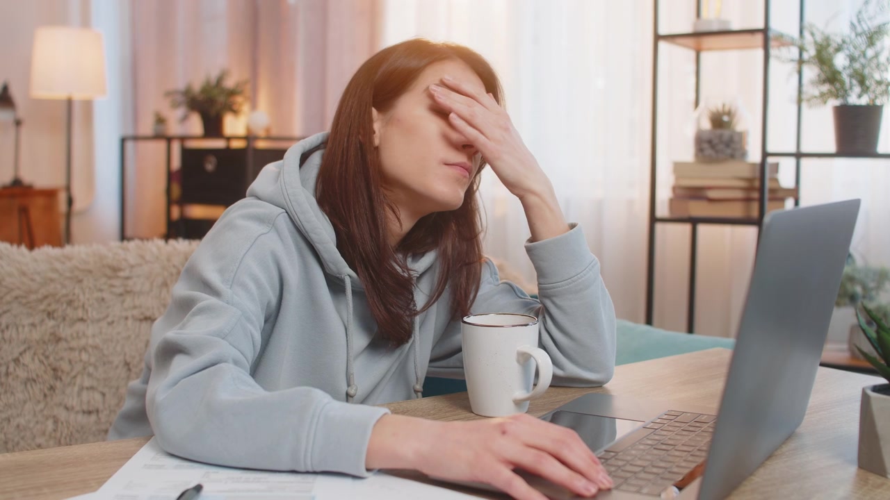 Giovane freelance stanca che usa il computer portatile al tavolo con documenti nel soggiorno di casa