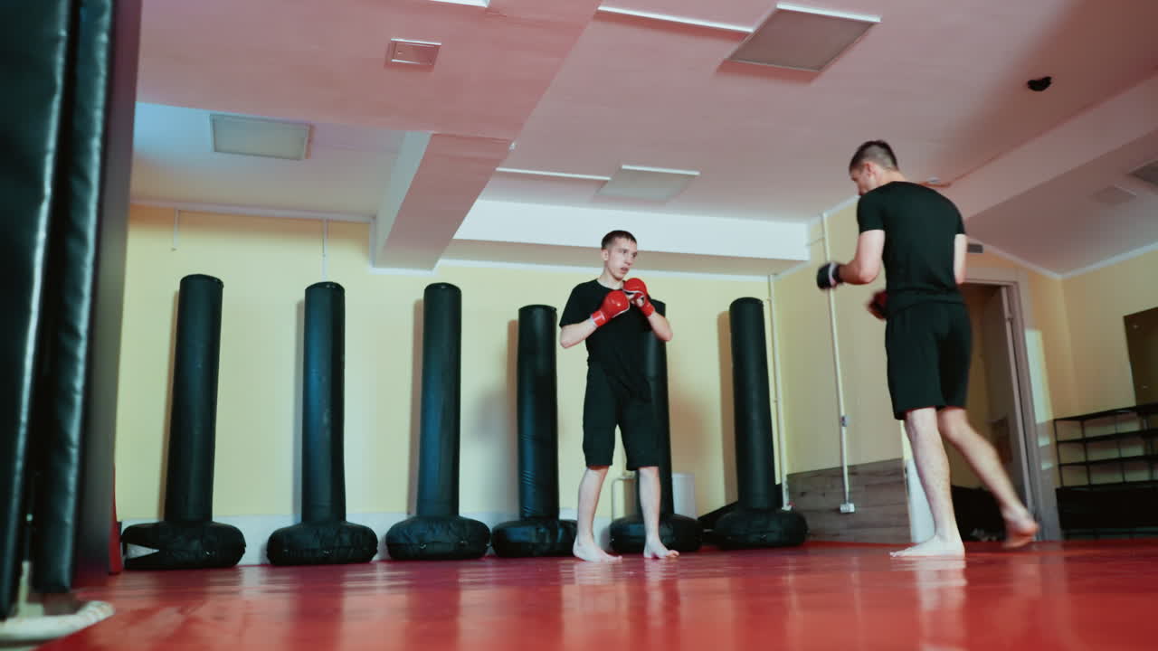 Athletes engaged in martial arts sparring session inside gym, wearing gloves and black outfits, combat sport moves with powerful high kick and defensive stance on red mat floor near punching bags indoors