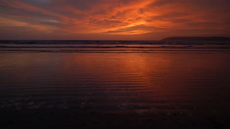 vista marina de las olas del océano rompiendo en la costa arenosa, durante una puesta de sol roja y naranja brillante, en una noche nublada