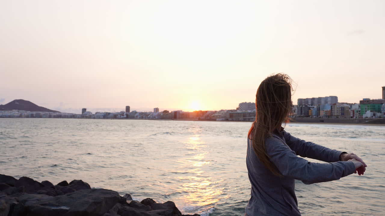 Young female doing yoga stretching on sandy Las Canteras beach, practicing mindful meditation during golden sunrise with ocean and volcanic landscape in background