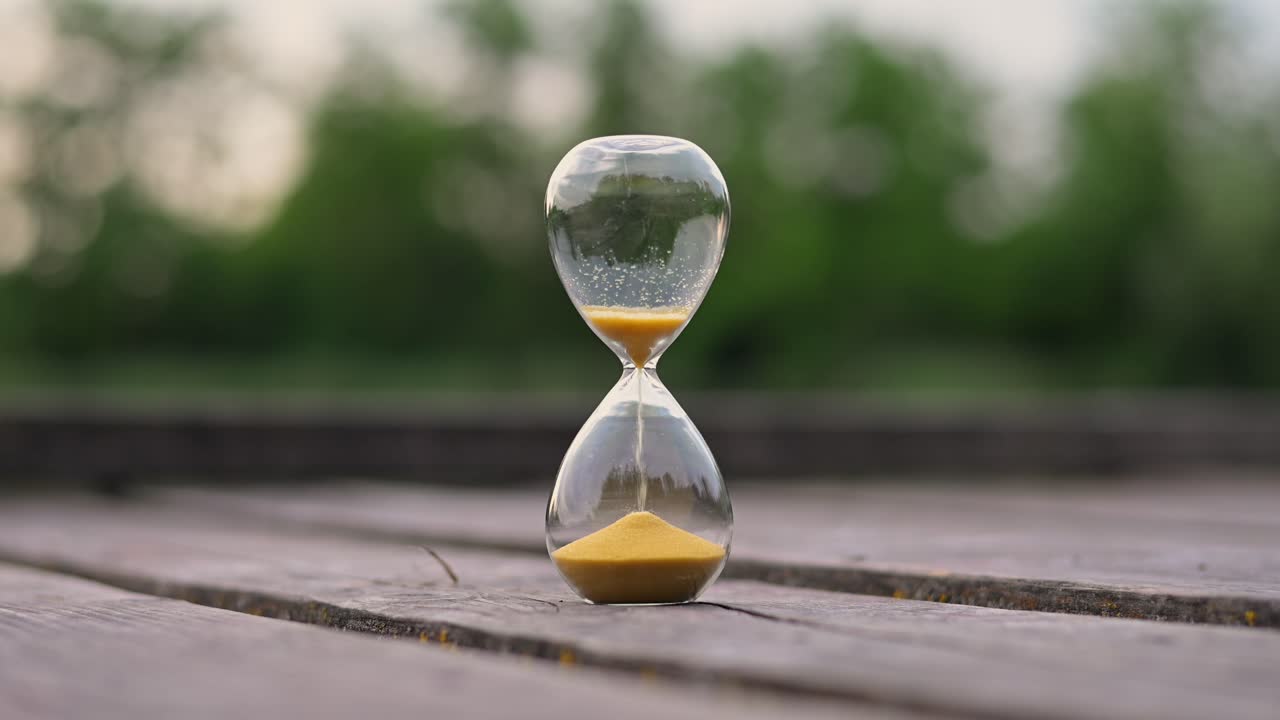A classic glass hourglass with golden sand is captured in mid-flow, placed on a wooden surface outdoors. The background features a softly blurred view of green trees, creating a serene atmosphere.