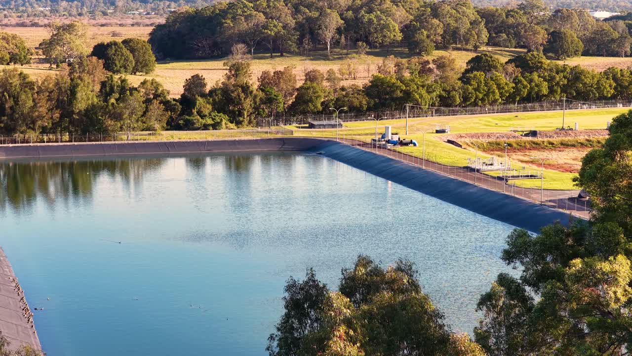 Aerial camera smoothly pans across a large wastewater treatment pond surrounded by greenery at sunset, highlighting reflective water and industrial infrastructure