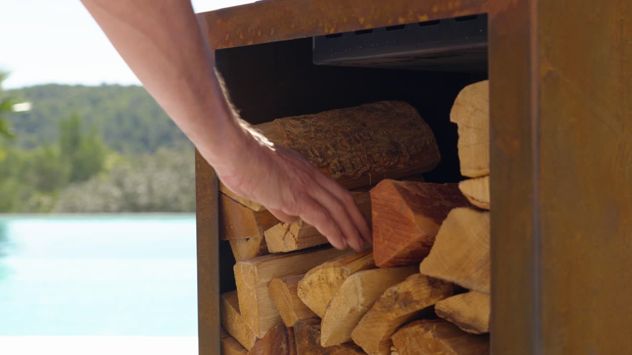 close-up of a pile of logs with a Caucasian man's arm reaching in to take a few pieces, blurred swimming pool in the background