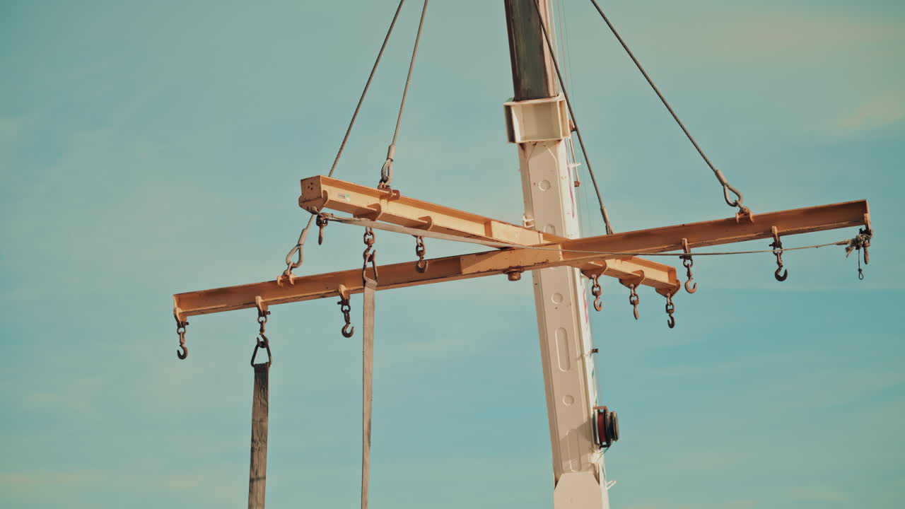 A yellow metal lifting beam with hooks, suspended by cables against a clear sky