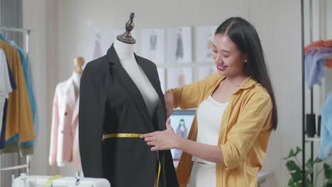 una diseñadora asiática sonriente midiendo una chaqueta de negocios en un maniquí mientras diseña ropa en el estudio