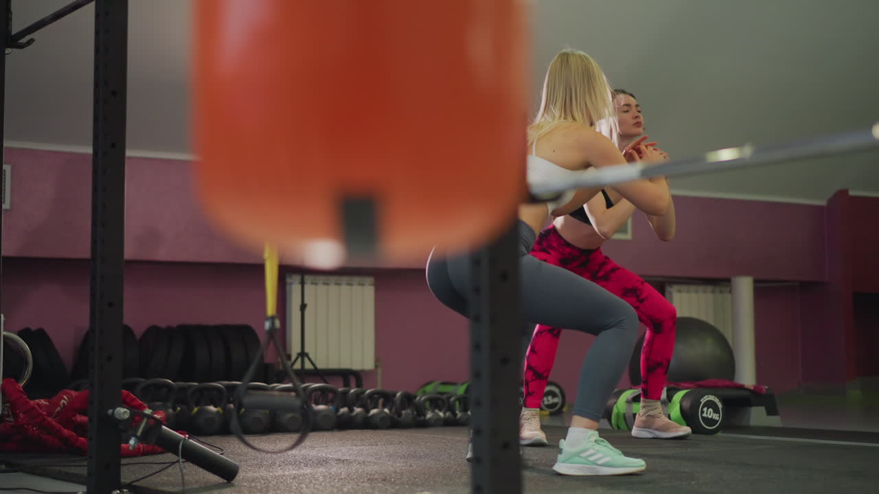 Squat con bilanciere in coppia, due atlete che lavorano insieme con il coach che le assiste e fornisce supporto in una palestra boutique, correzione ravvicinata di postura e profondità, illuminazione da studio brillante, rack